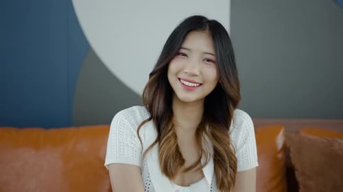 Smiling beautiful young asian woman looking at camera sit on sofa alone at home