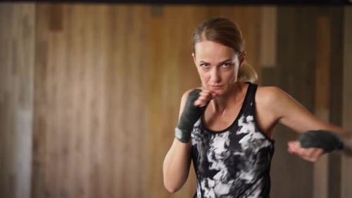 Boxerin macht Schlagbewegungen mit umwickelten Händen, trainiert alleine im Fitnessstudio und schaut in die Kamera