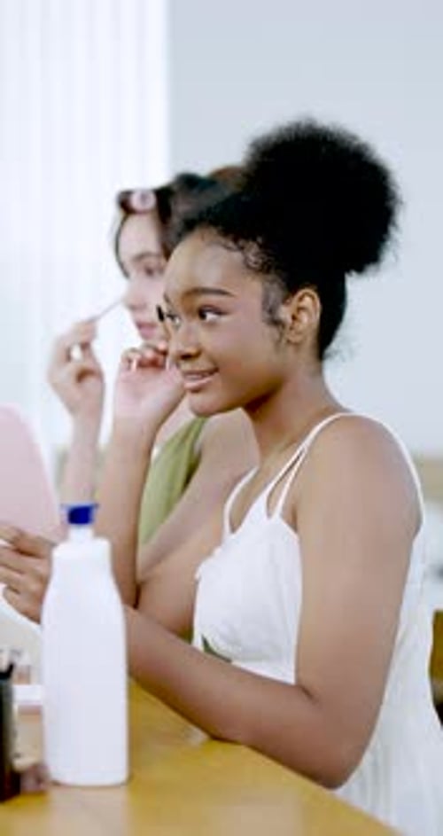 Afroamerikanisches Teenager-Mädchen beim Auftragen von Wimperntusche mit einem rosa Spiegel mit asiatischem Teenager-Mädchen