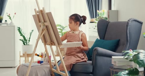 girl with apron sit on sofa chair in front of painting canvas with color palette use paint brush.