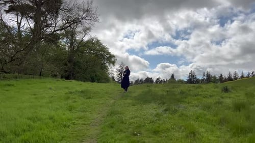 1 Girl with black dress walks towards trees green grass near meadow. View from behind turn around lo