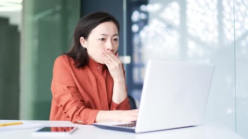 Traurige schockierte asiatische Geschäftsfrau liest schlechte Nachrichten auf einem Laptop, während sie am Arbeitsplatz sitzt