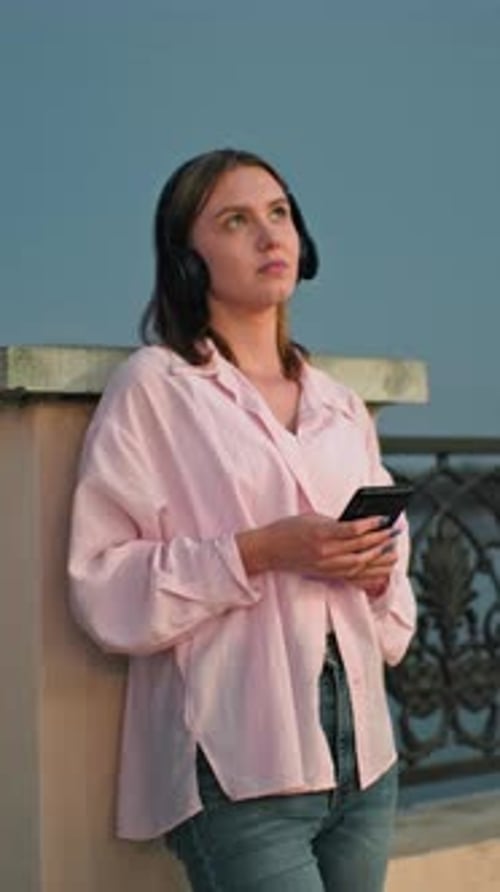 Woman in headphones leans against railing using phone