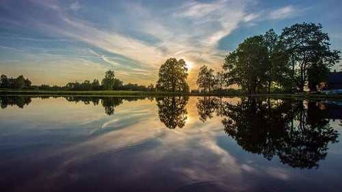 Timelapse shot of beautiful sunset over lake surrounded by trees with artistic clouds passing by.
