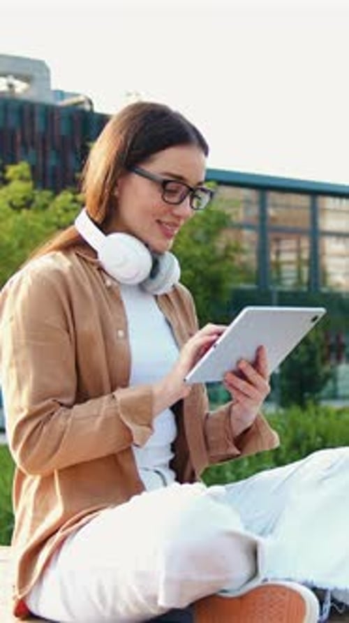Young Woman Sitting Crosslegged on Bench in Urban Outdoor Park Working on a Tablet While Wearing