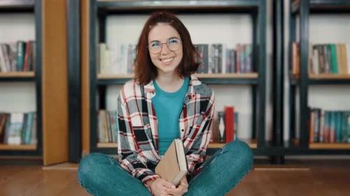 Portrait of Young Woman Student Reading a Book in the Library Sitting on the Floor Students in a