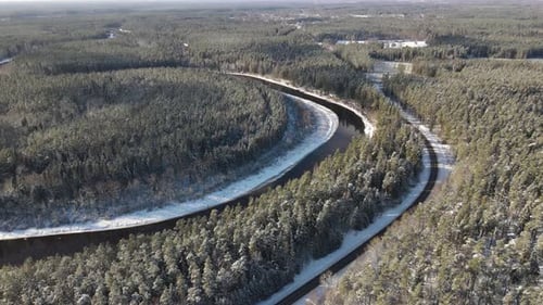 Aerial view of a winding river through a dense snow-covered forest in winter, with a curving road ru
