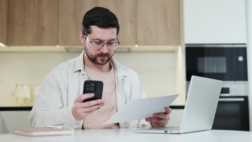 Thoughtful Male Comparing Documents From Smartphone with Original in Hands
