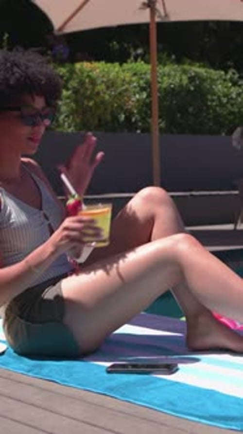 Reaching for cocktail, sun warming woman wearing striped tank top sipping on towel by pool phone