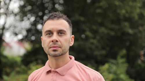 A Man in a Pink Polo Shirt Standing in Front of Trees on the Grass He Looks Focused Looking Away