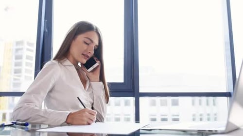 Talks by the phone. Young successful woman sits by table and works in the office