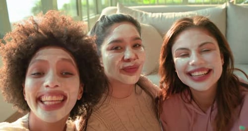 Amigas diversas estendendo o braço do smartphone capturando poses divertidas usando máscaras no sofá iluminado pelo sol