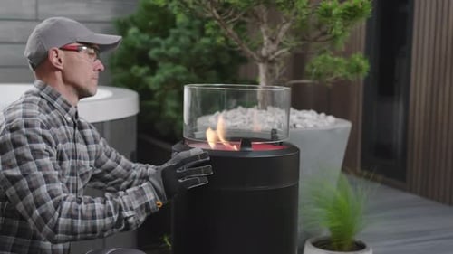 Man Sets up Outdoor Fire Pit Appliance in Backyard While Wearing Gloves and a Cap During Afternoon