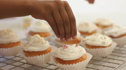 Hände, die nach links gehen, streuen goldene Streusel und rosafarbene Perlen auf die gefrosteten Cupcakes, um sie zu dekorieren.