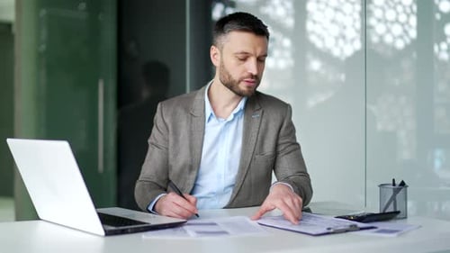 Busy businessman in a jacket is doing paperwork while sitting at a desk at workplace in business