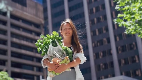 Mujer joven que lleva felizmente una bolsa de productos frescos en una ciudad
