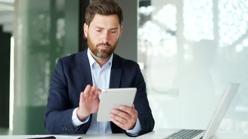 Businessman in formal suit is using digital tablet while sitting at workplace in business office.