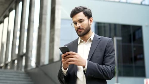 Portrait of happy businessman using mobile phone standing on street near office building. Smiling