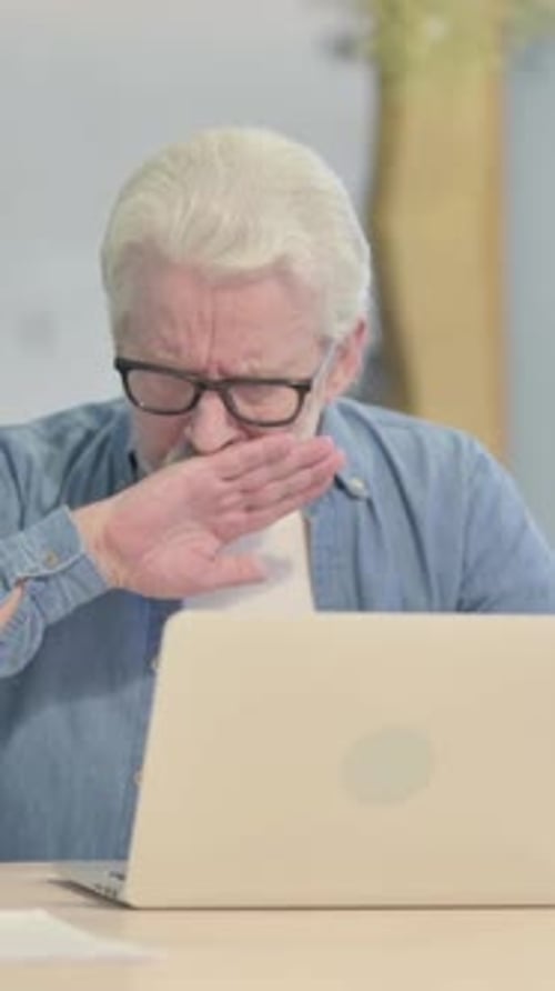 Senior Man Coughing While Using a Laptop at Desk