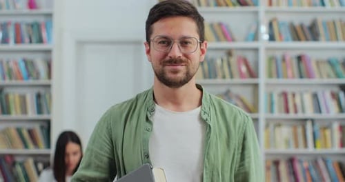 Close Up Portrait of Young Man in Glasses Holding Digital Tablet in Library Bookshelf Background