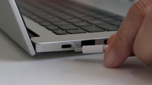 Hand plugging white USB-C charging cable into laptop, close up
