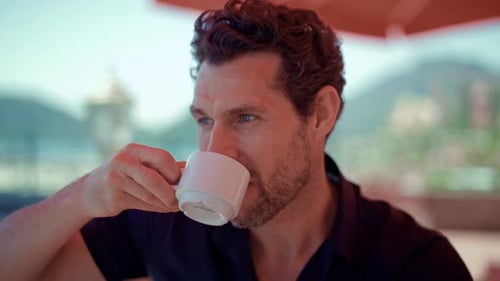 Smiling man enjoying a cup of coffee at a sunny seaside café with mountains and water view