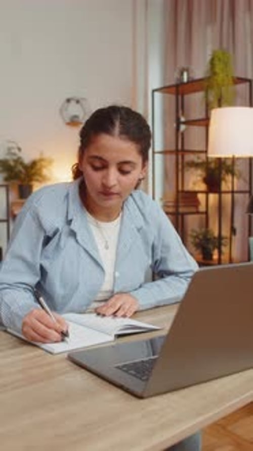 Young Woman Writing Down Notes While Attending Online Office Meeting on Laptop at Home Office Desk