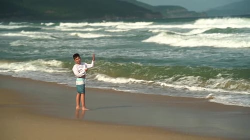 A Boy Stands on the Beach and Waves His Hand Against the Backdrop of Sea Waves