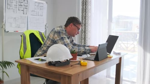 El coordinador del sitio caucásico trabaja en el escritorio con el casco y el chaleco de alta visibilidad con la computadora portátil abierta