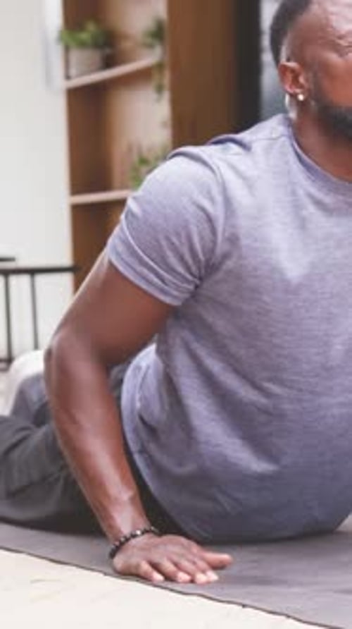 Vídeo vertical: Exhalando con fuerza, hombre haciendo presión sobre una colchoneta de yoga en casa para estirar la espalda en camiseta gris