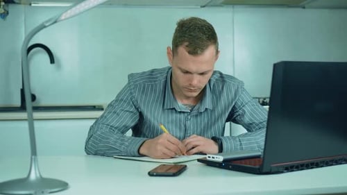 Focused Businessman Working on Laptop Computer in Home Office and Writing on Paper with Pen