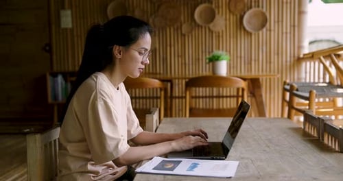 Asian woman adult sits at table working on laptop reviewing bar charts and pie graphs related to