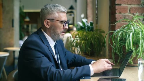 Mid-Aged Businessman Working on Laptop near Window in Modern Cafe