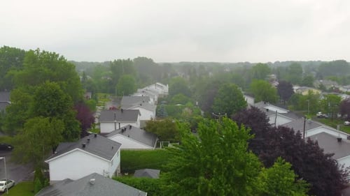 Drone view of white houses with gray roof, surrounded by trees under rainy Montreal sky