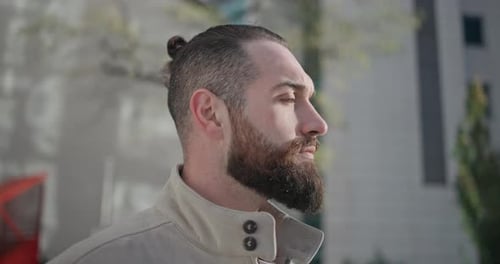A Young Man with Stylish Trendy Hairstyle and Beard in Casual Clothes Stands on the Street in the