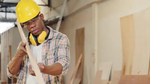 African American Carpenter Inspecting a Piece of Wood While Carrying a Wooden Plank in a Workshop