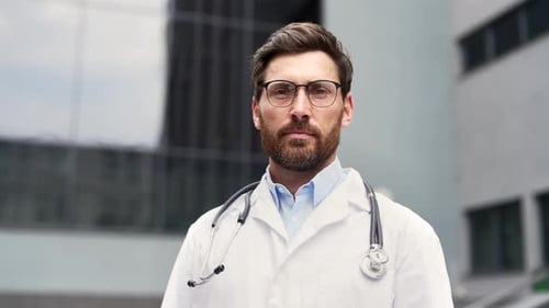Confident male doctor in white lab coat with stethoscope standing outside medical office.