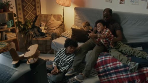 African American Man Cuddling Smiling Child on Sofa, Near Another Child on Floor