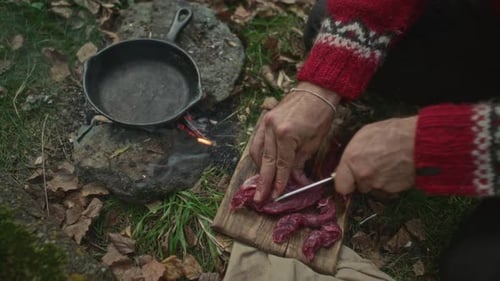 Hands of Man Cutting Raw Meat and Placing in Skillet over Campfire in Forest