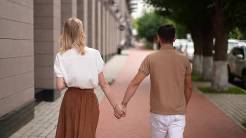 Rear View of Happy Young Couple Wearing Casual Clothes Walking Together on City Street and Holding