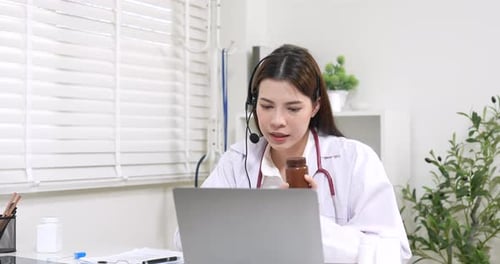 Asian woman doctor wearing headset consulting patient online using laptop, explaining medicine