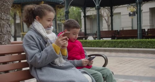 A Girl and a Boy are Sitting on a Bench in the Park Playing in Phone with Their Games Concept