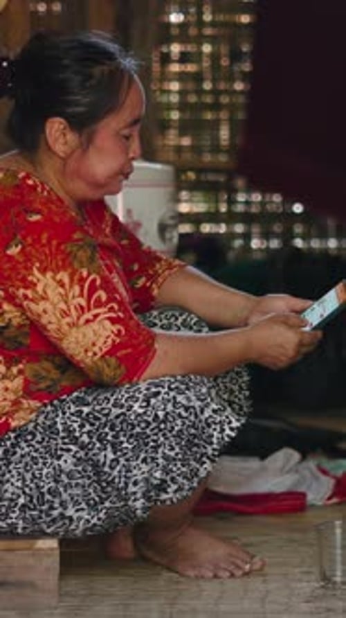 Mujer asiática de la tribu Lahu sentada descalza en su cabaña de madera consulta la pantalla de su teléfono inteligente