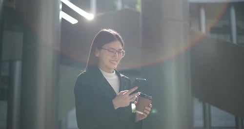 Smiling Woman Uses Phone and Holds Coffee Outdoors