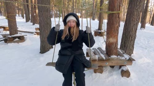 Happy Teen Girl is Riding on Swing Hanging on Rope Between Trees in Winter Park