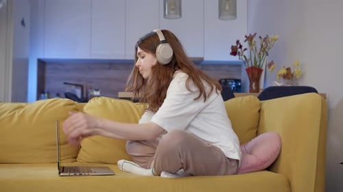 Junges Mädchen beendet die Arbeit am Computer auf gelbem Sofa, sie schließt den Laptop, entfernt ihre Kopfhörer und