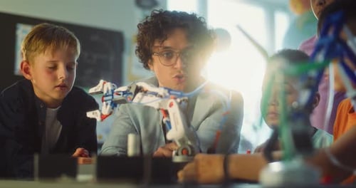 Team of Talented Kids Sitting at Table Studying Robotic Arm Model with Teacher in Primary School
