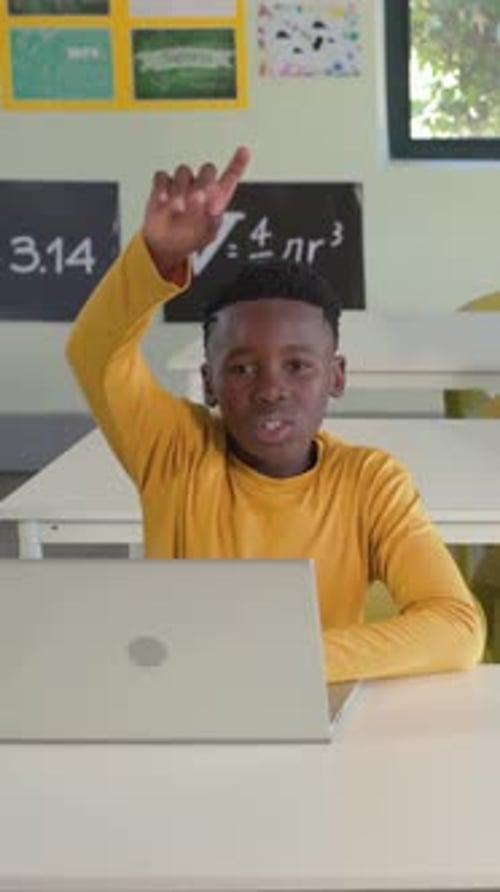 Vertical video: Pupil in tan shirt raising hand wanting to answer, typing on silver laptop in class