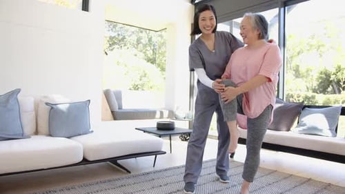 Physiotherapist assisting senior asian woman with leg exercise in living room
