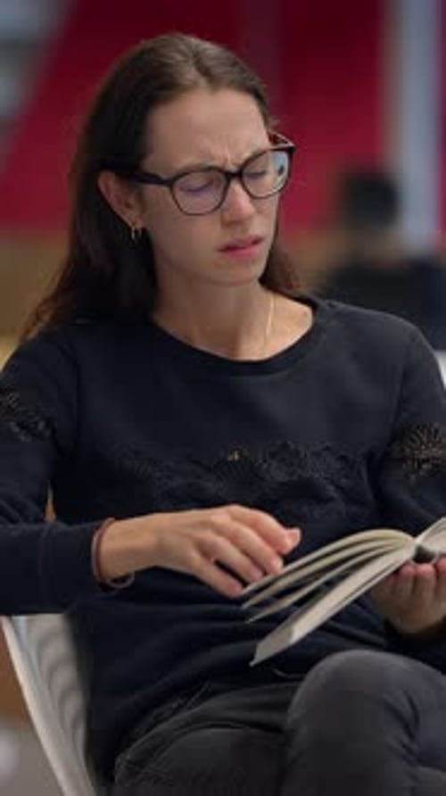 A Woman is Sitting Comfortably in a Modern Library Setting While Deeply Reading a Book